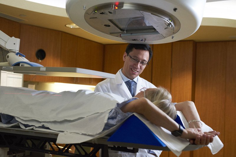Doctor in white coat standing next to treatment table with patient lying on her back underneath radiation devices.