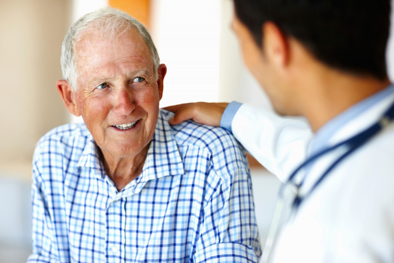 An older man in a checkered shirt smiles at his doctor, who has his hand on the man’s shoulder. 